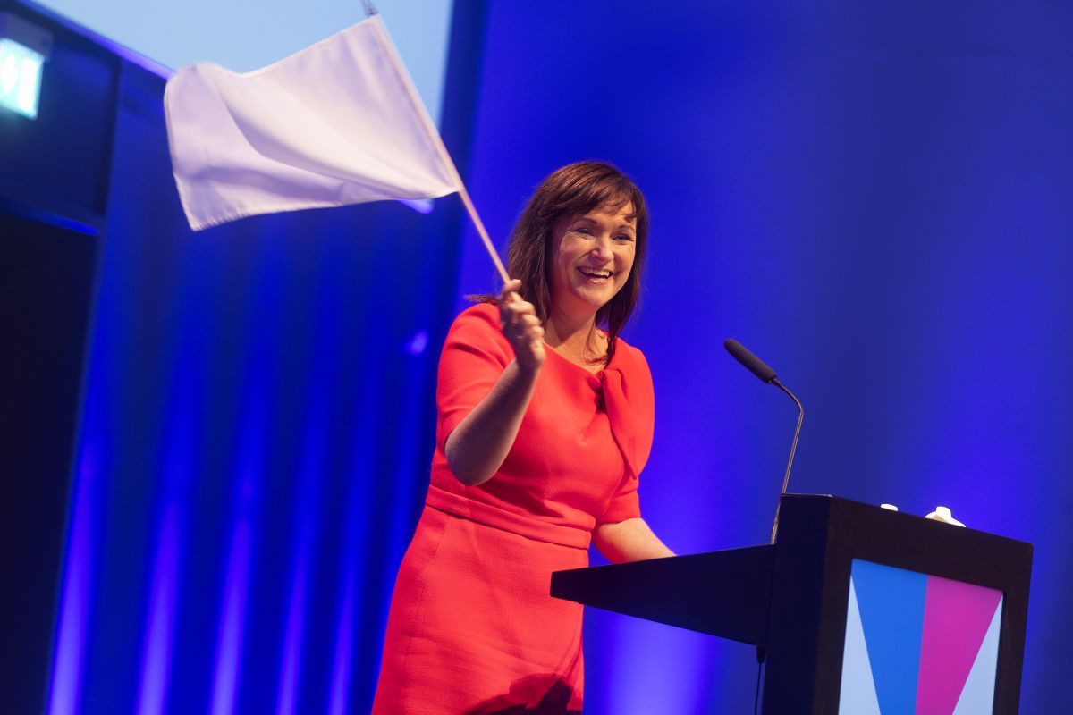 Sophie Løhde vifter smilende med et hvidt flag på talerstolen på Lægedages store scene. 