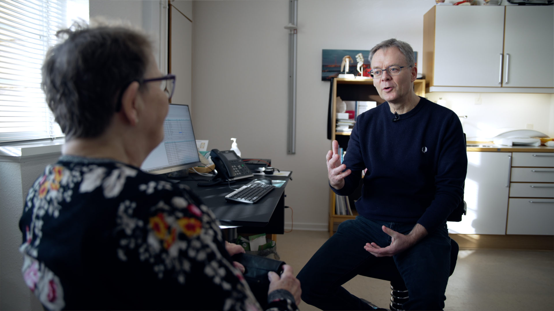 Praktiserende læge, mandlig, midaldrende, taler med patient, der sidder med ryggen til beskueren. 